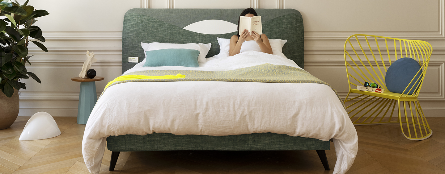Person lying on a bed with a green Treca headboard in a room with a yellow chair and plant.