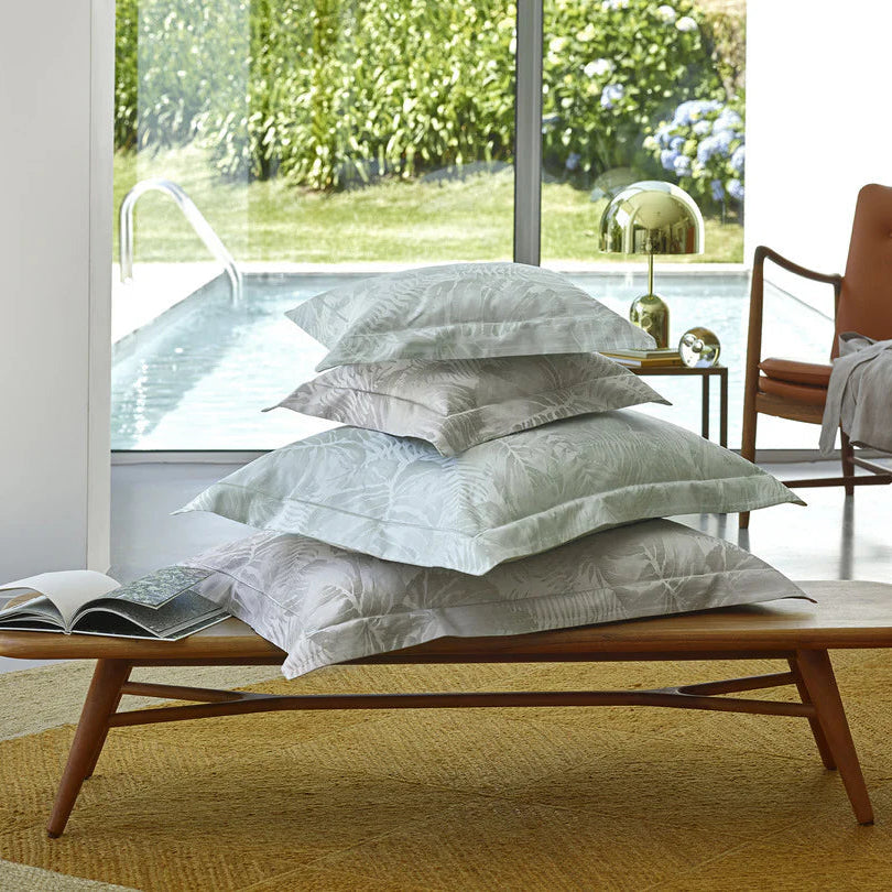 Stack of pillows with Copacabana jade pillowcase sham on a wooden bench in a bright room with large windows.